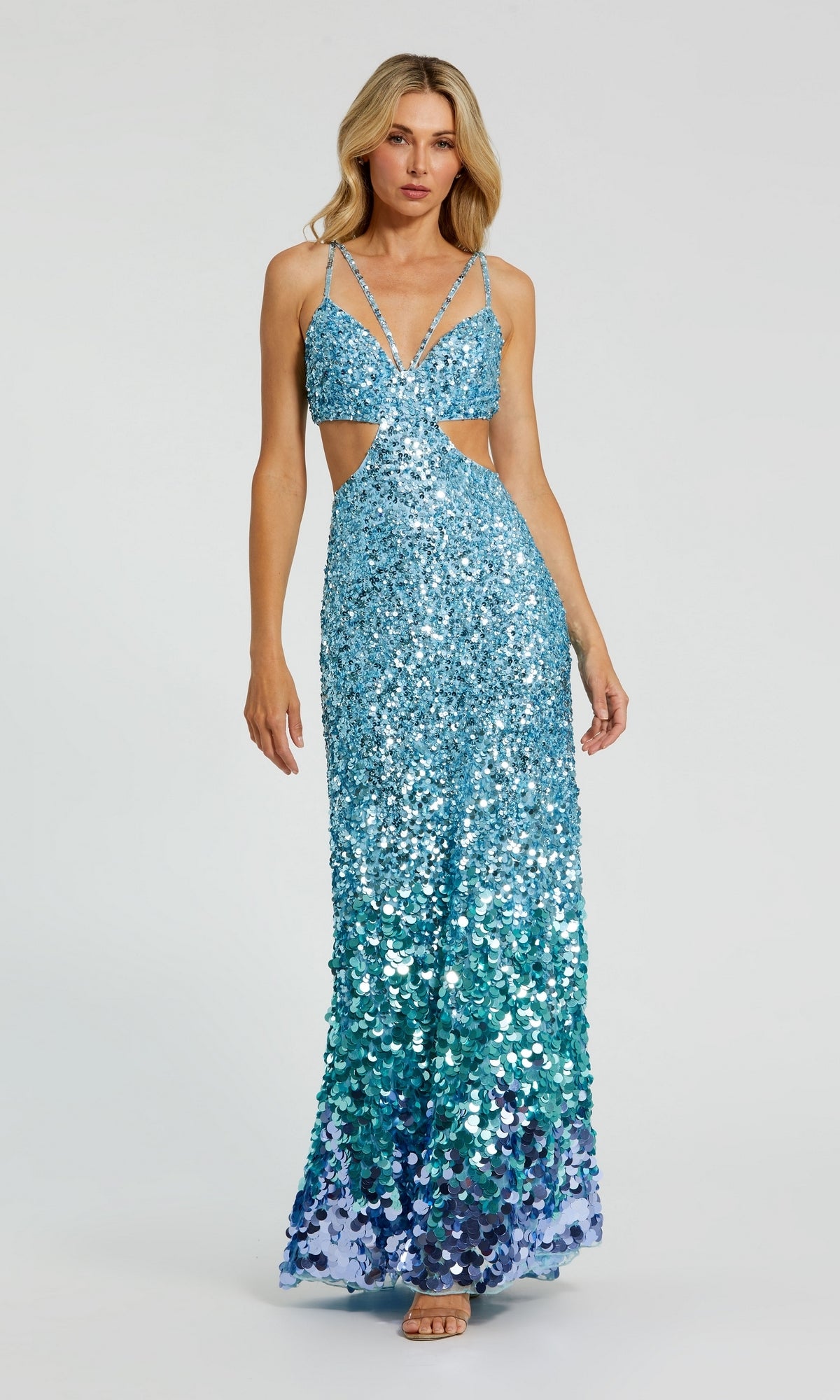 A woman models the Long Formal Dress 6077 by Mac Duggal—blue-green sequins, cut-out sides, and thin straps—while standing against a plain light gray background.